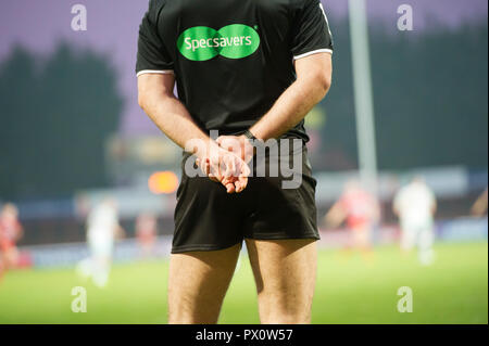 Arbitro indossando top nero e corti sul passo di rugby durante il gioco specsavers logo le mani incrociate dietro la schiena i giocatori al di fuori della messa a fuoco Foto Stock