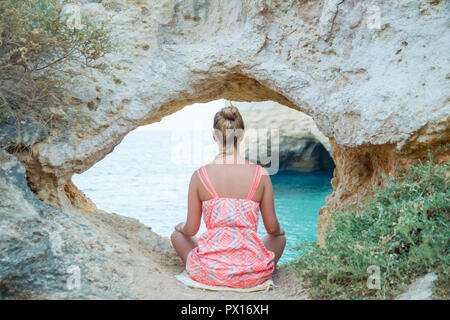 Giovane donna pratica di meditazione durante il tramonto sotto una pietra calcarea naturale formazione di arco da una laguna turchese del Algarves, Portogallo. Foto Stock