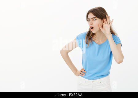 Voi dite che cosa. Ritratto di incuriosito interessati carino donna in blu t-shirt, piegando verso la telecamera, tenendo la mano vicino a orecchio, intercettazioni gossip o overhearing interessante conversazione oltre il muro grigio Foto Stock