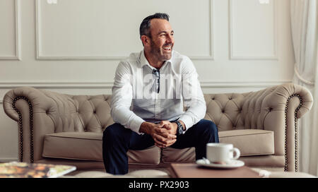 Ritratto di Allegro imprenditore seduti sul divano nella camera di albergo e guardando lontano da ridere. CEO di stare in un lussuoso hotel room in viaggio d'affari. Foto Stock