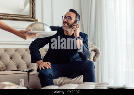 Imprenditore maturo seduto sul divano parlando al telefono cellulare in camera in hotel con un cameriere che serve cibo. Servizio in camera Servizio cameriere che serve cibo di un imprenditore Foto Stock