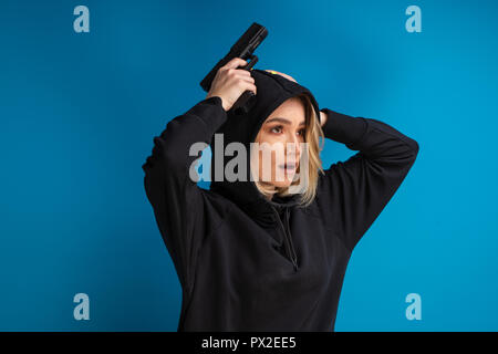 Ritratto di ragazza con cappuccio guardando scioccato tenendo le mani con la pistola fino sulla testa. Colpo contro sfondo blu Foto Stock