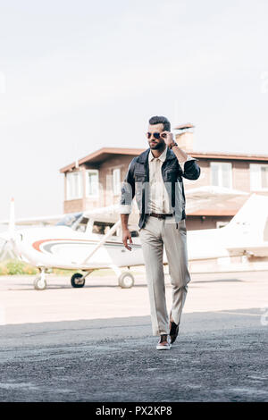 Bello elegante uomo in giacca di pelle e occhiali da sole passeggiando nei pressi di aeroplano Foto Stock