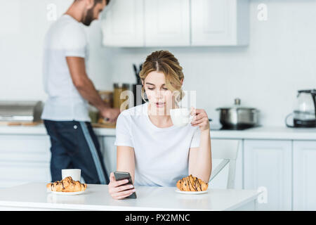 Giovane donna a bere caffè e utilizza lo smartphone mentre fidanzato cooking per colazione dietro Foto Stock