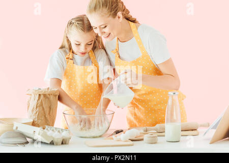 Felice madre versando il latte nella pasta durante la figlia isolato per impastare in rosa Foto Stock