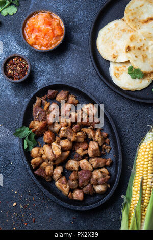 Cibo colombiano. Maiale fritto CHICHARRON, AREPAS colombiano e salsa di pomodoro. Vista dall'alto. Sfondo nero Foto Stock