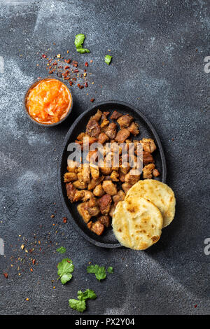 Cibo colombiano. Maiale fritto CHICHARRON, AREPAS colombiano e salsa di pomodoro. Vista dall'alto. Sfondo nero. Foto Stock