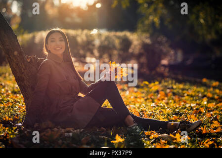 Donna durante l'autunno, il concetto di stile di vita. Un asiatico giovane femmina adulta è godendo il sole durante la caduta, in un parco con caduto foglie gialle. Foto Stock