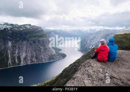 Trolltunga - più spettacolari attrazioni in Norvegia. Coppia di viaggiatori si siede sulla roccia e guarda al lago Ringedalsvatnet Foto Stock