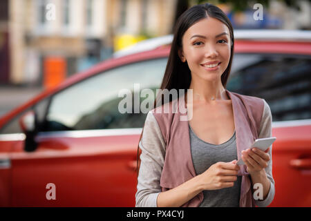 Una giovane bella donna asiatica in posa con il suo smart phone nella parte anteriore di una macchina nuova di zecca. Comunicare su smart phone applicazioni sulle auto Foto Stock