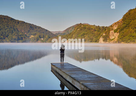 Una donna sul suo proprio nelle prime ore del mattino in piedi sulla estremità di un pontile Llyn Gwynant guardando la campagna circostante, Snowdonia, Wales, Regno Unito Foto Stock