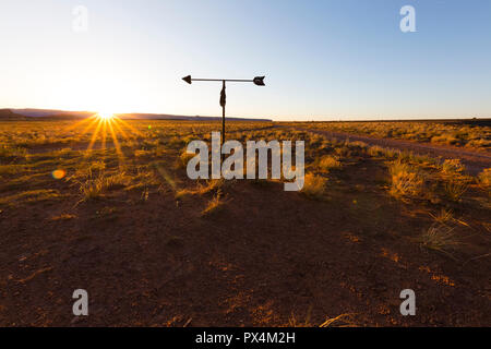 Shash Dine Navajo campeggio, AZ, Stati Uniti d'America. Il sole tocca l'orizzonte su una banderuola segnavento e il deserto di macchia. Foto Stock