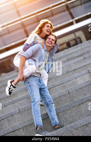 Uomo di dare la sua bella ragazza una piggy back a street sorridente a vicenda in una giornata di sole. Foto Stock