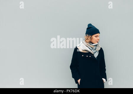Femmina cappotto indossa, beanie cappello e sciarpa, guardando verso il basso, copia dello spazio. Giovane donna in piedi all'aperto in climi freddi blu sul muro solido background. Foto Stock
