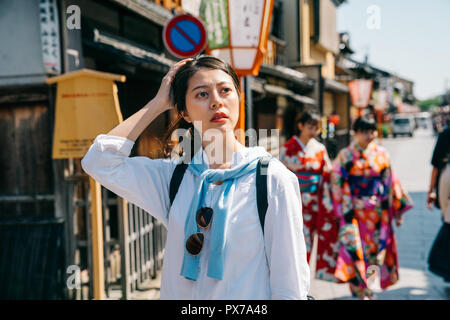 Elegante traveler è di toccare la sua testa mentre pensando che il modo giusto di hostel in Giappone. due giovani ragazze in kimono camminare in background. Vite giapponese Foto Stock