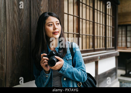 Un elegante fotografo asiatici basandosi sulle pareti in legno e guardando gli altri con la fotocamera sulla sua mano. Giappone viaggi vacanze estate ragazza godendo Foto Stock