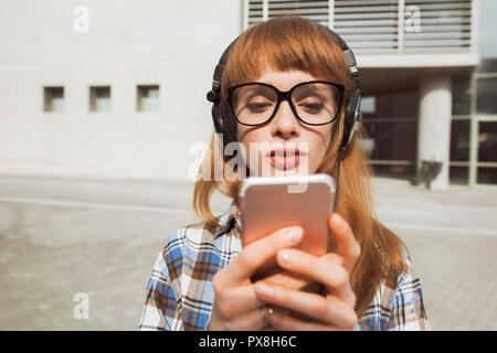 Un giovane hipster donna con i capelli rossi, utilizza un'applicazione nel suo smart phone dispositivo per inviare un messaggio di testo e ascoltare musica Foto Stock