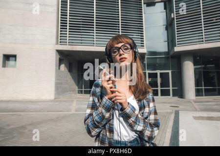 Un giovane hipster donna con i capelli rossi, utilizza un'applicazione nel suo smart phone dispositivo per inviare un messaggio di testo e ascoltare musica Foto Stock