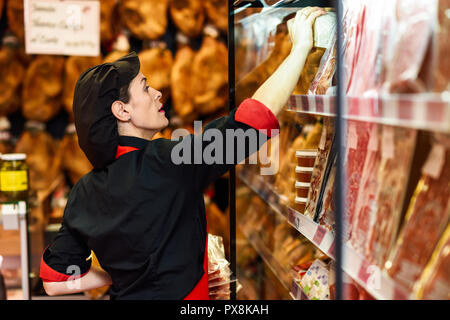 Ritratto di lavoratore di sesso femminile che prendendo prodotti di macelleria. Espositore refrigerato per pacchetti di salsicce, prosciutto e formaggio Foto Stock