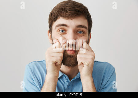 Close-up ritratto di cupo sconvolto maschio barbuto, schiacciando le guance con le mani e guardando la telecamera, essendo in cattivo umore mentre in piedi su sfondo grigio. Guy ha guadagnato peso extra durante le vacanze invernali Foto Stock