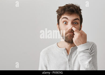 Funny barbuto ragazzo con capelli disordinati e cattive abitudini, fissando la telecamera con faccia stupido, picking nel naso con il dito indice e piedi su sfondo grigio. Coppia geek non conosce l'igiene Foto Stock