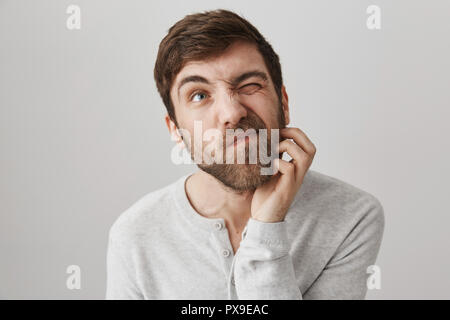 Ritratto di disordine maturo ragazzo caucasico guardando verso l'alto e graffiare la barba, pensando a qualcosa o di pianificazione viaggio futuro, in piedi contro lo sfondo grigio. L uomo decide se egli vuole davvero andare fuori Foto Stock