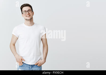 Il piacere di successo europeo guy celebrando la firma di buon affare con gli investitori, erge orgogliosamente sorridente largamente, ridere, tenendo le mani nelle tasche, indossare occhiali e t-shirt oltre il muro grigio Foto Stock