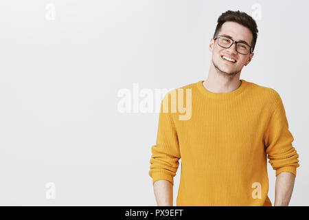 Vita-up shot di felice e soddisfatto bel giovane in bicchieri e maglione giallo testa tiltante, sorridere e ridere come cercando cordiale alla fotocamera sul lato destro dello spazio di copia su sfondo grigio Foto Stock