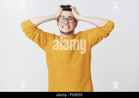 Uomo di ripartizione di essere pressurizzato e addolorato tira i capelli fuori di testa dei denti di aggraffaggio alla ricerca intensa e briga indignazione permanente e alimentato fino a maglione giallo e bicchieri oltre il muro grigio Foto Stock