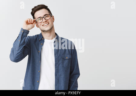 Ritratto di Nizza moderno e bello studente maschio in bicchieri e maglietta blu toccando il telaio del eyewear sorridente cordiale e felice e soddisfatto con il team produttivo lavoro sul progetto oltre il muro grigio Foto Stock