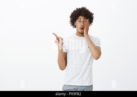 Ritratto di scioccato impressionato bello di carnagione scura maschio con acconciatura afro e baffi, piega le labbra con bocca aperta, tenendo palm sulla guancia, puntando in alto a sinistra sulla parete grigia Foto Stock