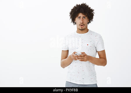 Oh no, perché. Ritratto di sconvolgere cupo e confuso carino ispanica modello maschile in t-shirt bianca con lo smartphone nelle mani, accigliata dalla tristezza, dimenticare la password, impossibile effettuare il login tramite il dispositivo Foto Stock