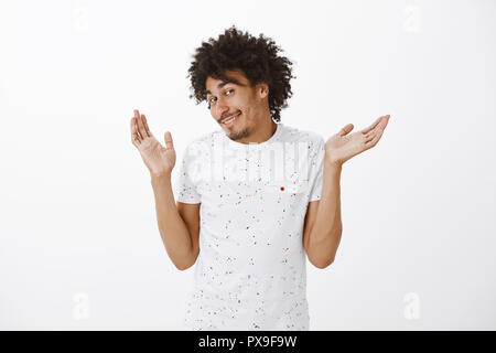 Non è un mio problema. Ritratto di spensierata e indifferente carino felice ragazzo con capelli ricci e i baffi, facendo alzare la spalla sollevata con palme, sorridente largamente, essendo clueless e ignora Foto Stock