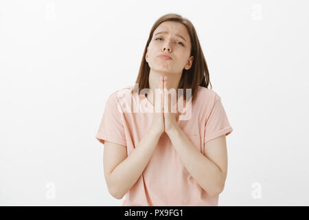 Si prega di fare di me favore, mendicante. Ritratto di carino donna europea in rosa t-shirt, busbana francese e accigliata guardando con espressione triste in telecamera, tenendo le mani a pregare chiedendo perdono Foto Stock