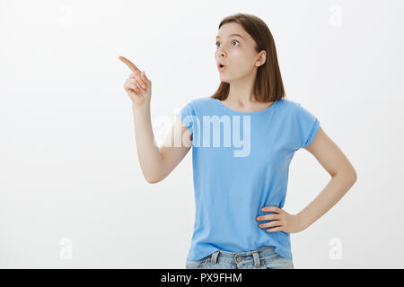 Colpito bella ragazza in blu t-shirt, tenendo la mano sul hip, dicendo wow mentre cercano e puntamento nell'angolo superiore sinistro, essendo scioccato e lieta di vedere incredibili e cosa incredibile Foto Stock