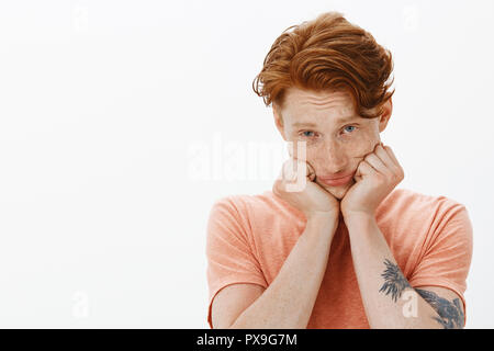 Carino redhead uomo europeo con lentiggini sul viso, testa pendente su mani e guardando con cupo sconvolto l'espressione in telecamera, con la voglia di gelato o dolci, essendo moody e infelice, posa su parete grigia Foto Stock