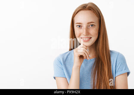 Curiose e interessate attraente redhead modello femminile con graziosi lentiggini sorridente con gioia mentre listenign storia interessante tenendo la mano sul mento che esprimono intrighi e gioia sulla parete grigia Foto Stock