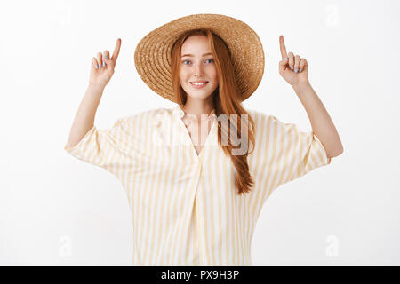 Ragazza vuoi andare a fare escursioni a piedi in montagna mentre si è in viaggio con il mio ragazzo in piedi nella trendy camicetta giallo e l'estate cappello di paglia i bracci di sollevamento verso l'alto e sorridente gioiosamente alla fotocamera sulla parete grigia Foto Stock