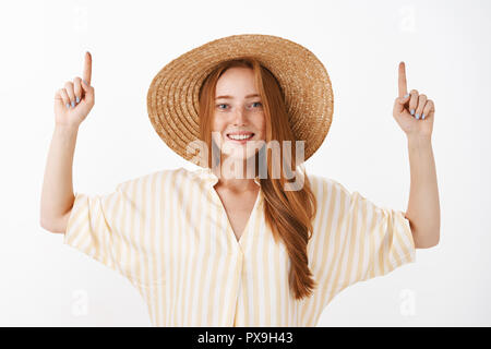 Vita-up shot di socievole e comunicativa bella elegante redhead donna in giallo Elegante camicetta e cappello di paglia i bracci di sollevamento verso l'alto mentre sorridente largamente in telecamera su uno sfondo grigio Foto Stock