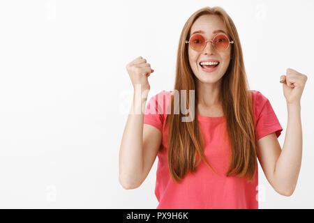 Redhead donna il tifo per il mio ragazzo mentre si esegue sul palco alzando i pugni di alta in modo favorevole spostare sorridente urlare con gioia mantenere alto il tentativo di accrescere la fiducia in piedi su sfondo bianco Foto Stock