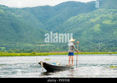 Giovani locali pescatore birmano indossando una maglietta verificato tornando a casa dopo la pesca, in piedi sul bordo di un sottile tall barca a remi, indietro, Lago Inle Foto Stock