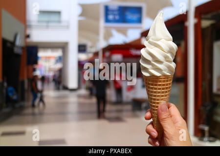 Donna di mano che tiene la vaniglia latte soft servire il gelato in cono di luce del sole, con sfocato shopping mall in background Foto Stock