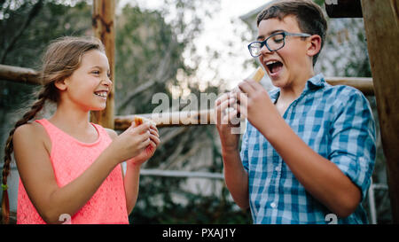 Sorridente un ragazzo e una ragazza mangiando panini in piedi in un parco. Contenti i bambini godendo il loro picnic mangiando snack all'esterno. Foto Stock