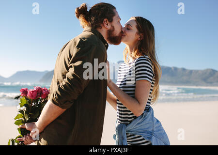 Uomo con sorpresa bouquet di rose per la bella giovane donna sulla spiaggia. Amare giovane baciare sulla spiaggia. Foto Stock
