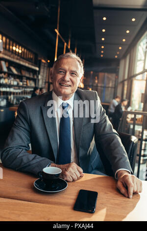 Ritratto di fiducia senior imprenditore seduto alla caffetteria moderna con tazza di caffè e telefono cellulare sul tavolo. Imprenditore maschio di relax presso il caffè sho Foto Stock