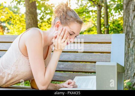 Una giovane donna è sdraiato su una panchina nel parco in un parco e la lettura concentrata in un libro Foto Stock