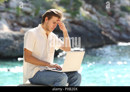 Uomo preoccupato leggendo le cattive notizie online in un computer portatile in vacanza sulla spiaggia Foto Stock