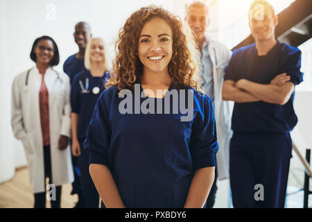 Sorridente giovane medico donna in piedi in un corridoio di ospedale con un gruppo eterogeneo di personale medico in piedi dietro di lei Foto Stock