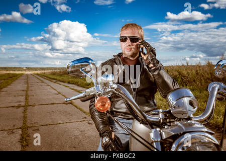 Biker parlando su uno smartphone. Biker uomo che indossa una giacca di pelle e occhiali da sole seduto sulla sua moto. Foto Stock