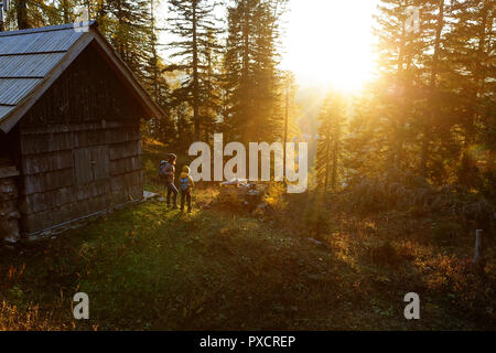 Matjer e figlio in piedi da una capanna in legno su Krstenica prato in autunno al tramonto luce dorata con larici intorno a loro, sulle Alpi Giulie, Slovenia Foto Stock